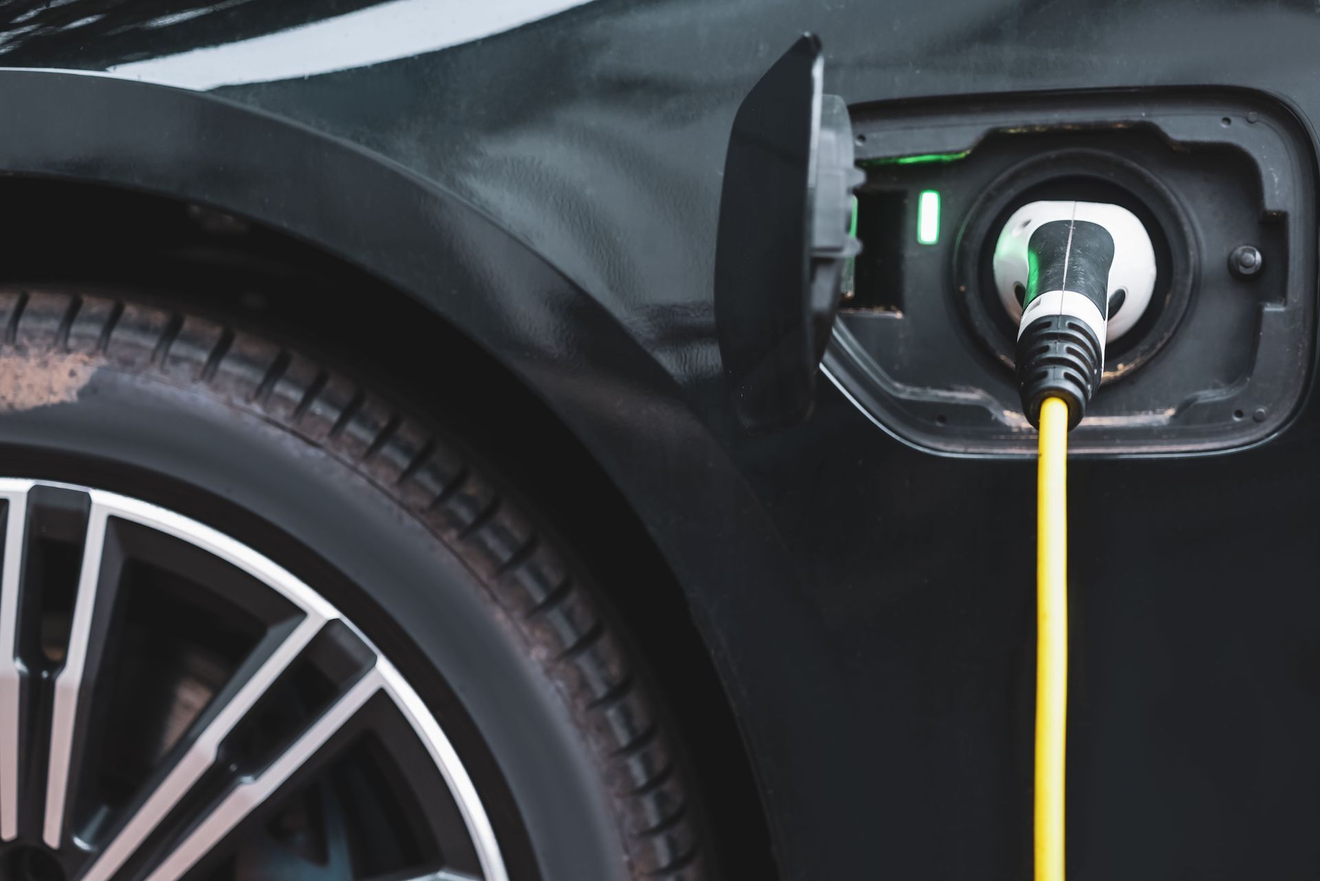 A close-up of a black electric car charging, with a yellow cable plugged into the side port showing a green light.