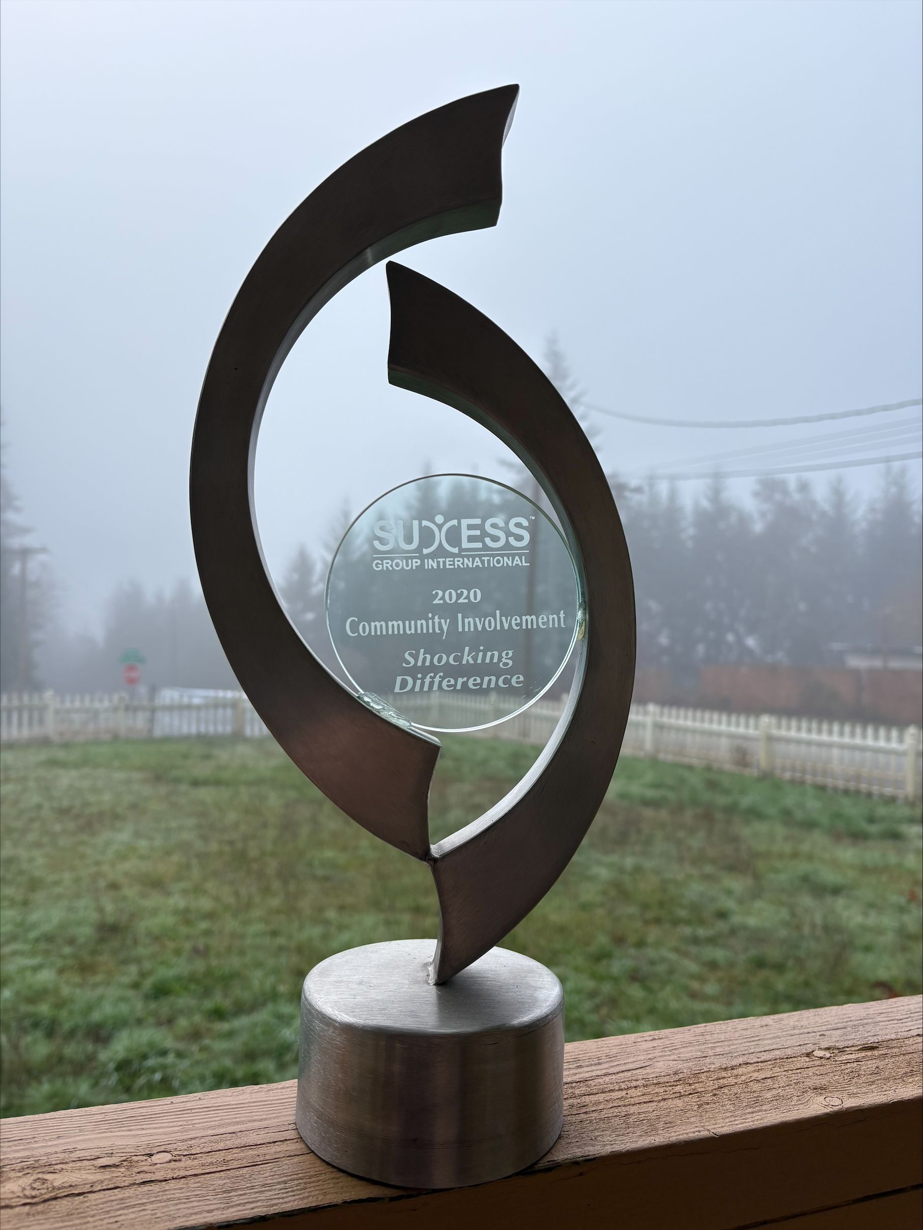 A metallic award trophy with a circular center panel, standing on a wooden surface against a foggy, outdoor background.