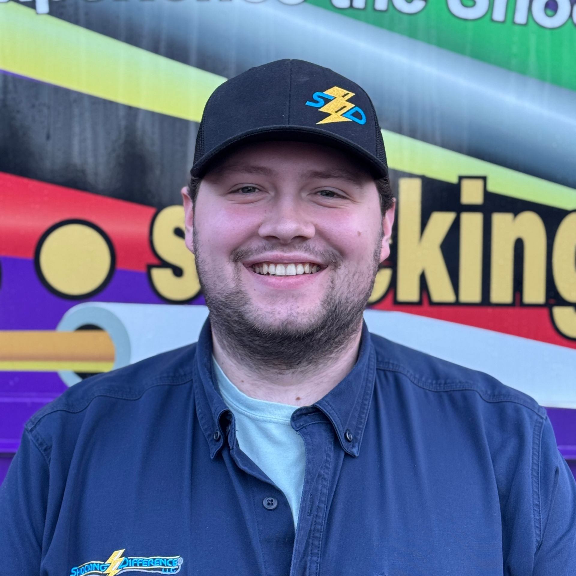 A smiling individual wearing a black cap with a logo and a navy blue button-down shirt stands in front of a colorful wall.