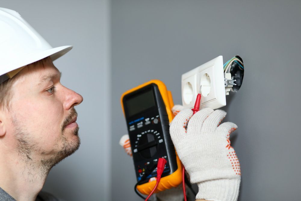 Electrician testing a white wall outlet with a multimeter, wearing a hard hat and gloves.