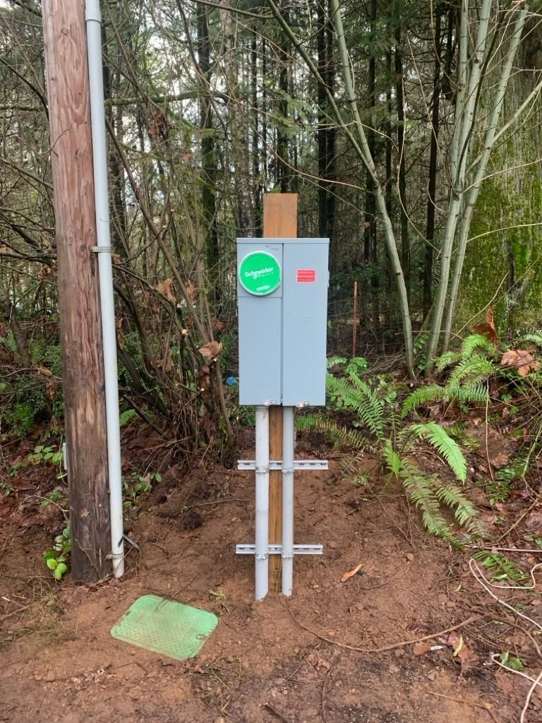 Gray utility box on metal posts in a wooded area beside a dirt path and green marker.
