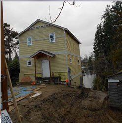A two-story, yellow-sided house with a small front porch under construction, situated on an unfinished dirt lot.