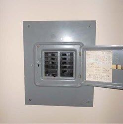 A grey electrical service panel with its door open, mounted on a plain wall, showing multiple circuit breakers inside.