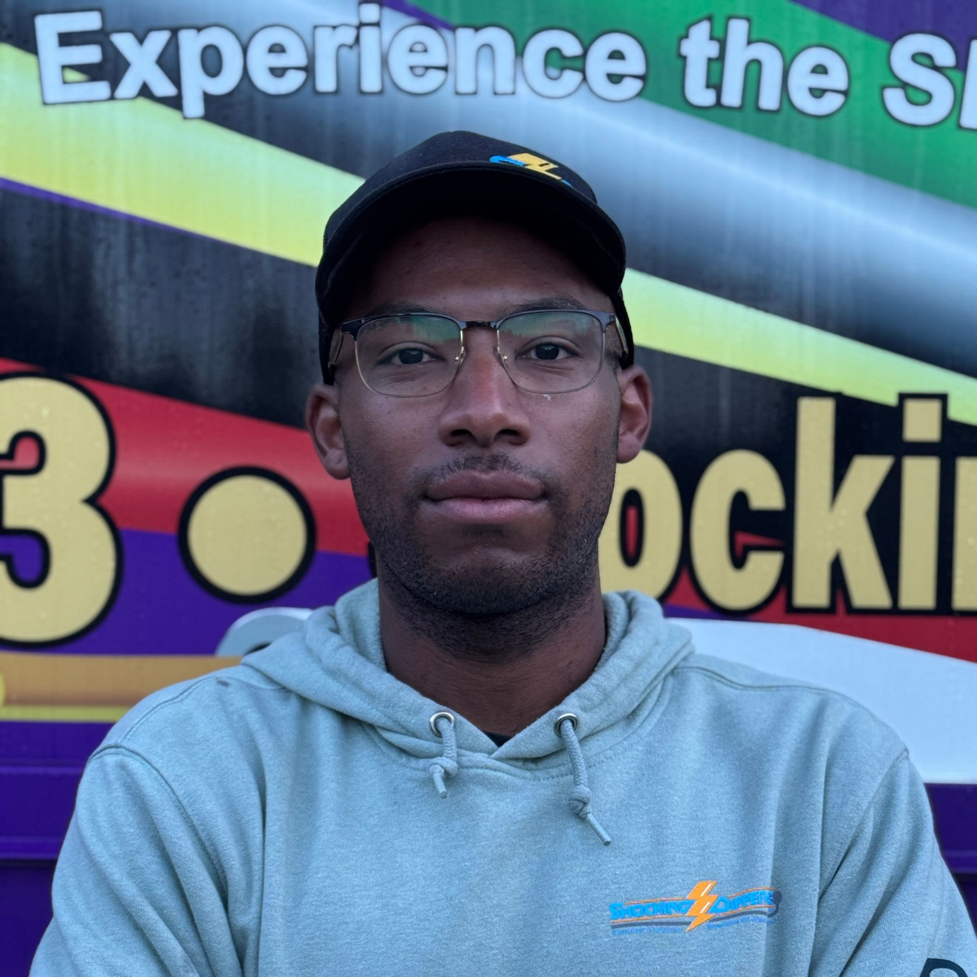 A person with glasses and a cap wears a light gray hoodie in front of a banner with text and colorful stripes.