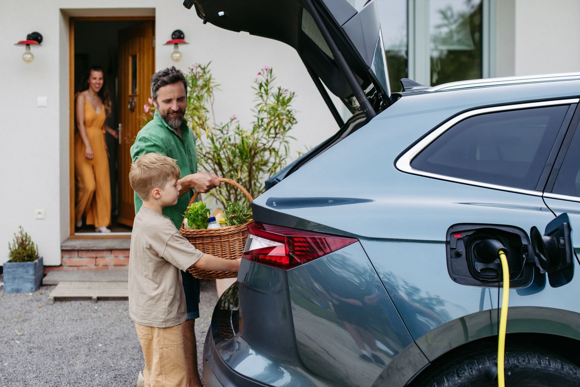 A person and child place a basket of groceries into the open trunk of an electric car charging at a home entrance.