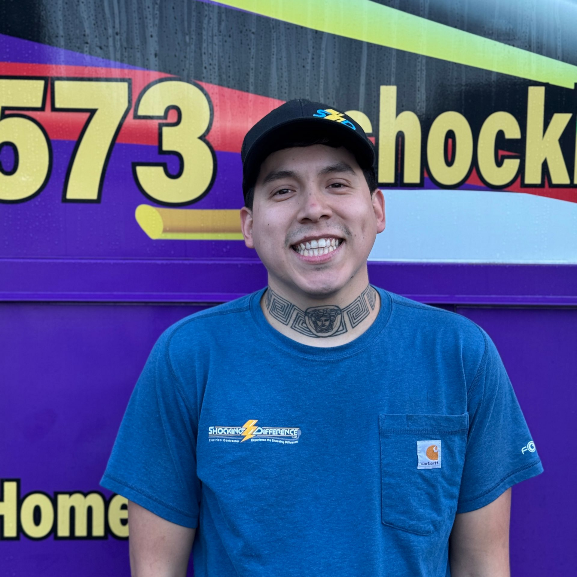 A smiling person in a blue t-shirt and black cap standing in front of a purple vehicle with yellow text.