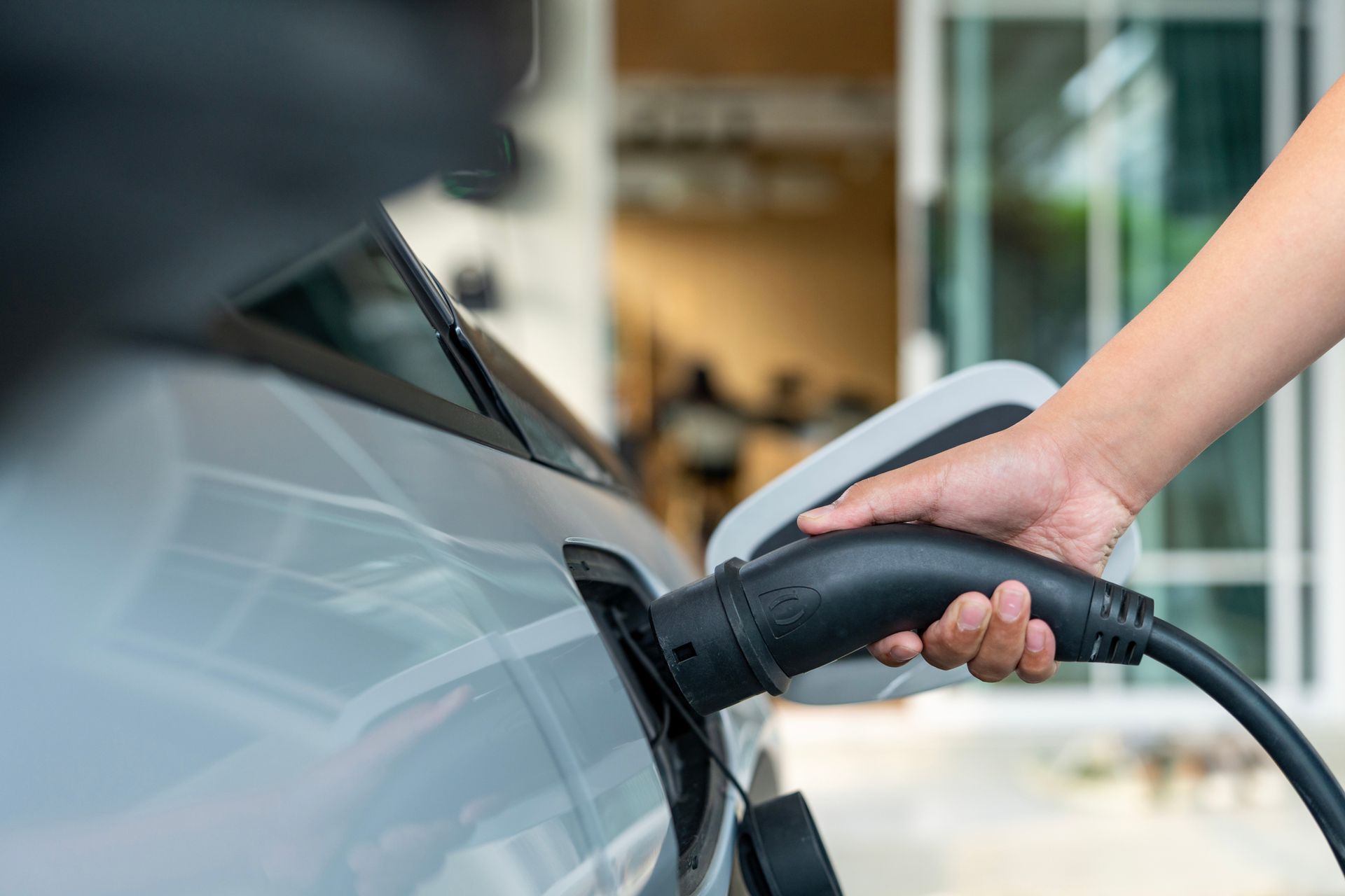 A hand holds a black charging cable and inserts it into the port of a silver electric vehicle.