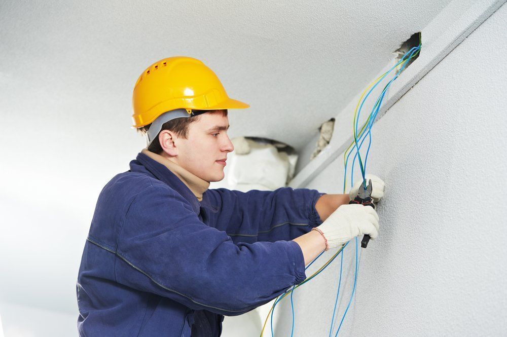 A construction worker in a yellow hard hat and blue work jacket strips electrical wires from a ceiling opening.