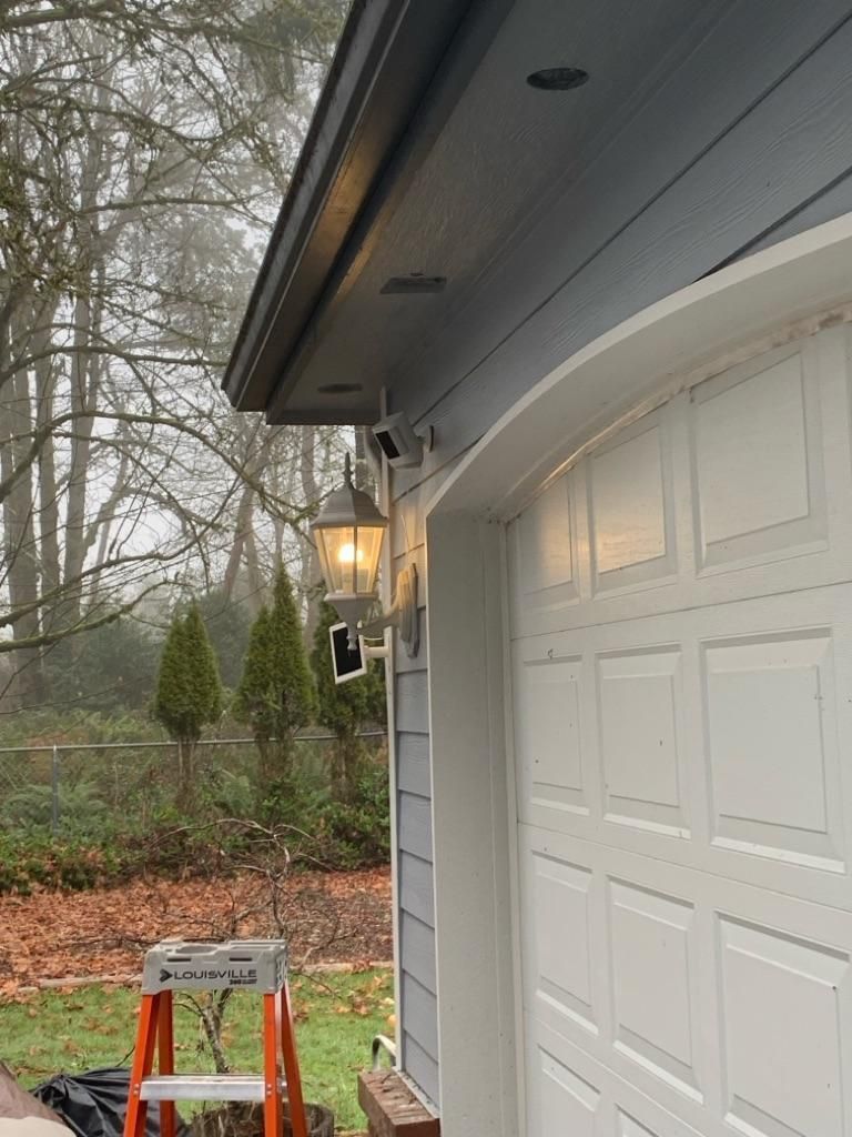 White garage door beside lit porch lantern and red stepladder near a wooded driveway in misty weather
