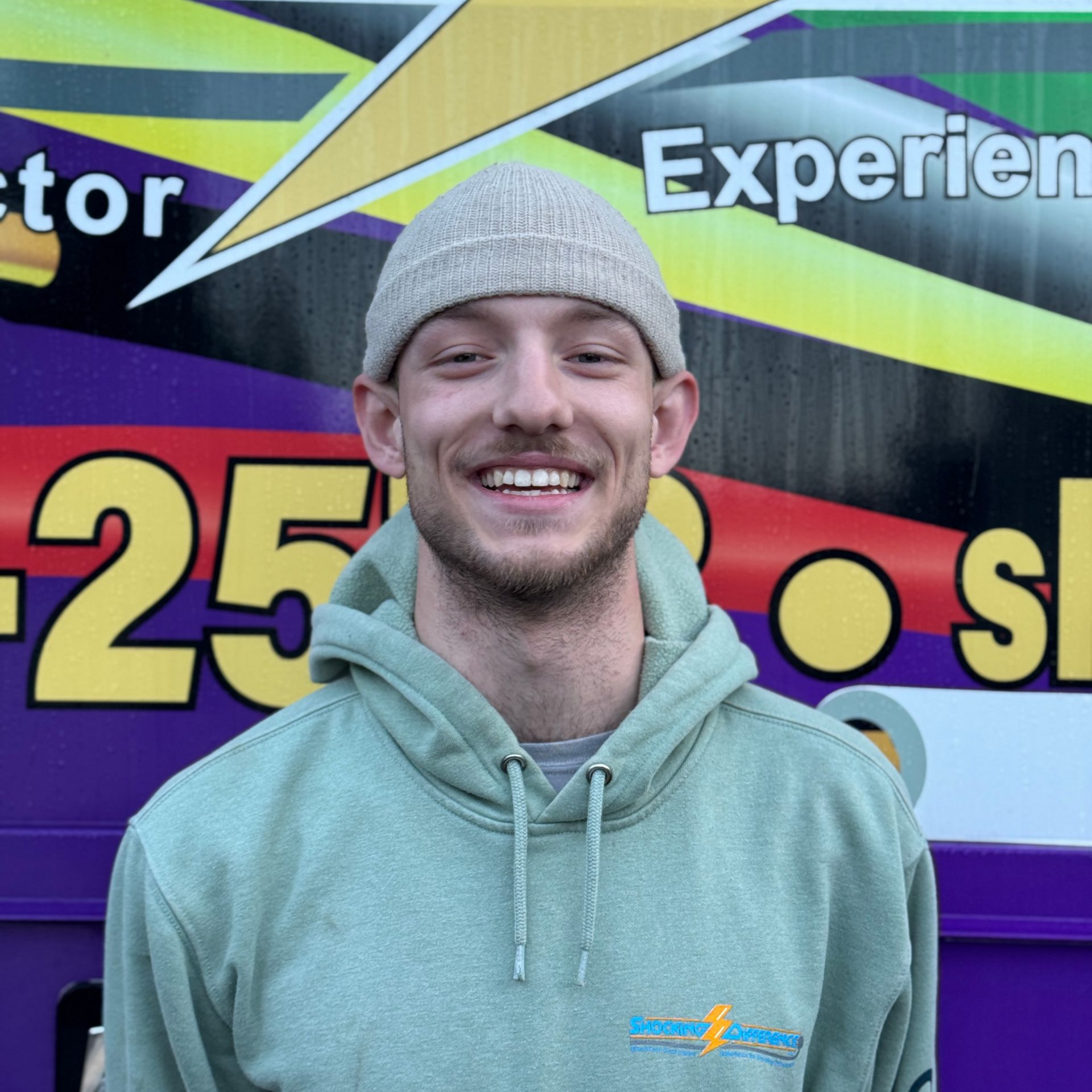 A person in a light green hoodie and beige beanie smiling in front of a purple, yellow, and black graphic background.