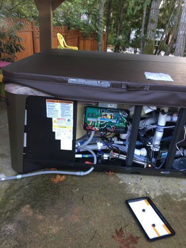 A hot tub with its side panel removed, revealing the interior plumbing, electrical box, and machinery on a patio.