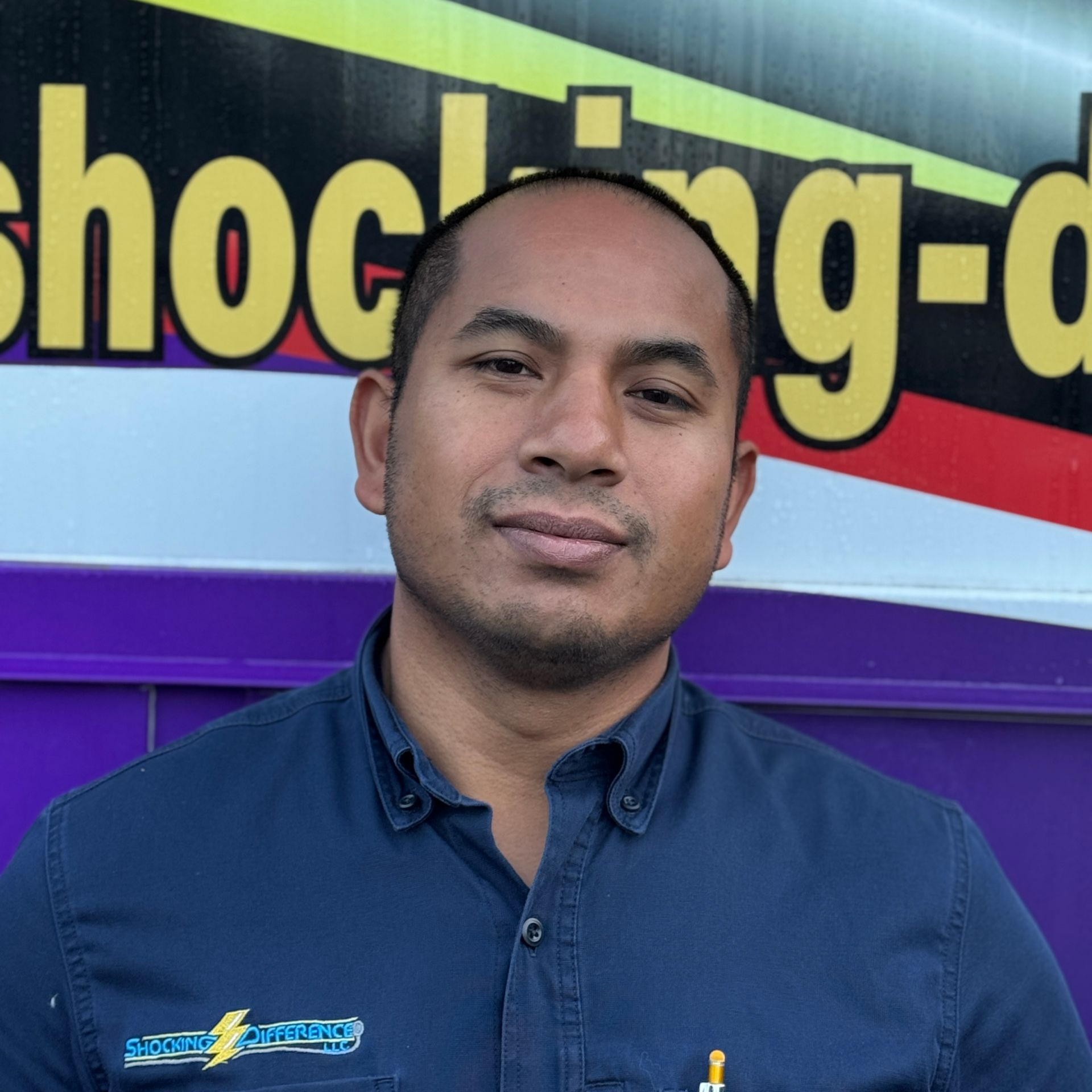 A person in a dark blue button-down shirt stands in front of a purple and yellow background with partial text.