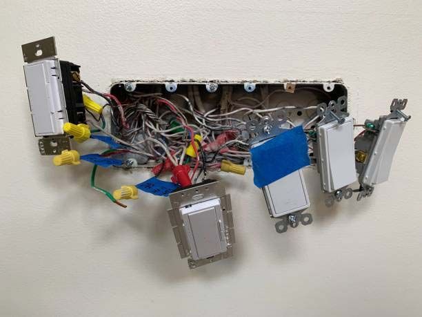 Open electrical junction box with tangled wires and several wall switches mounted on a beige wall