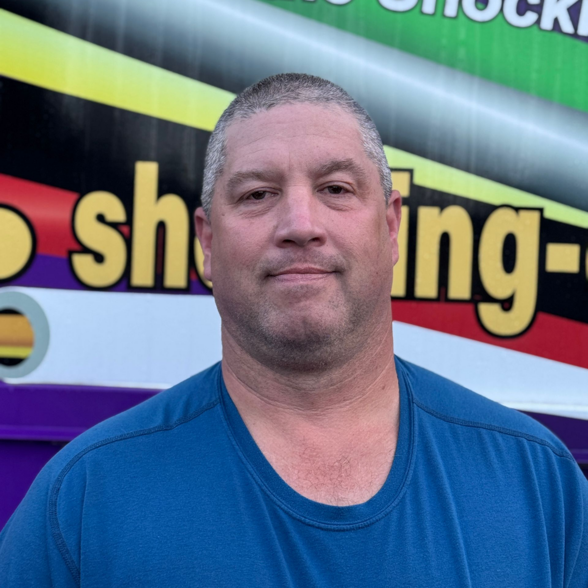 A person in a blue t-shirt stands in front of a colorful, patterned sign with yellow lettering.