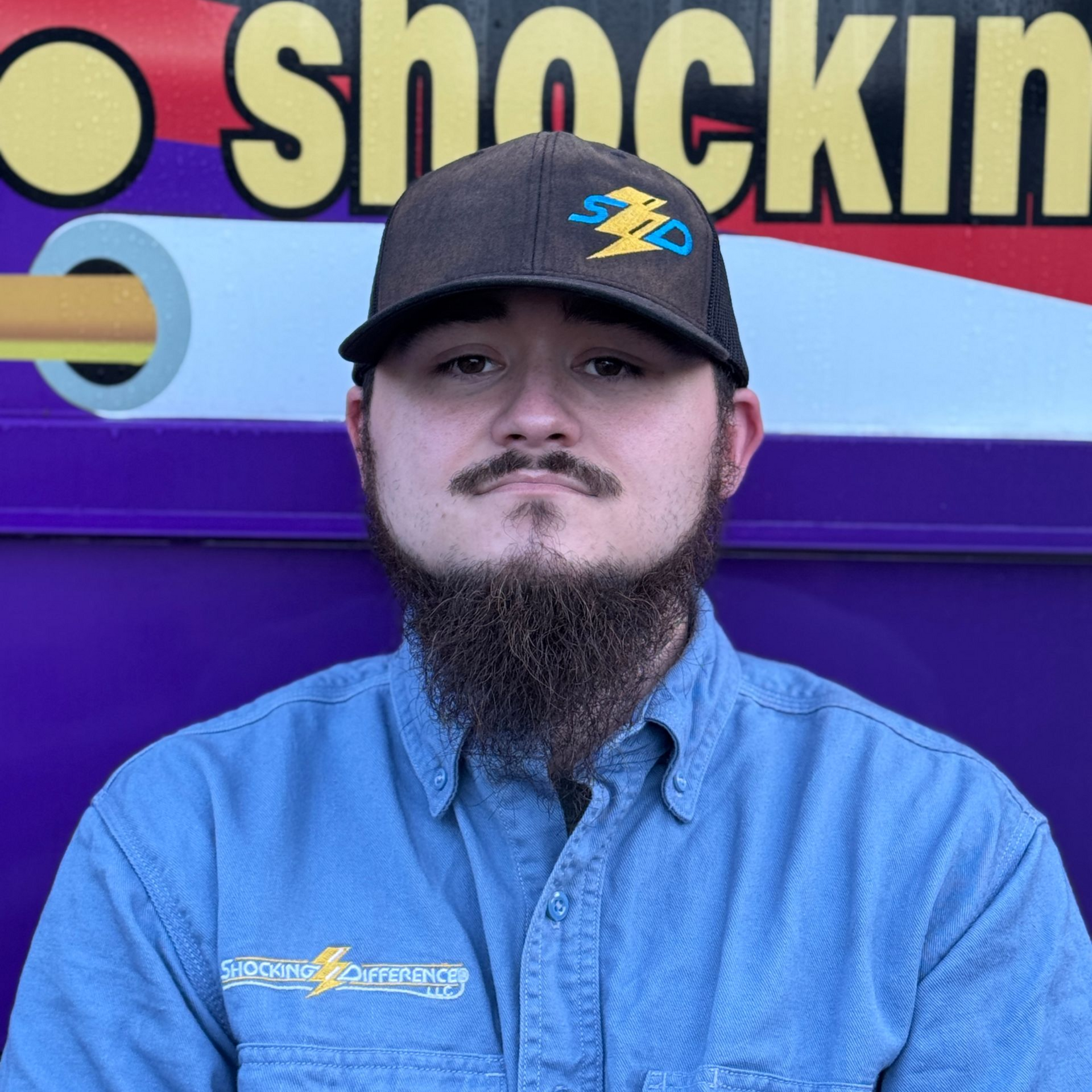 A person wearing a blue shirt and dark baseball cap with a lightning bolt logo, posed in front of a purple branded vehicle.