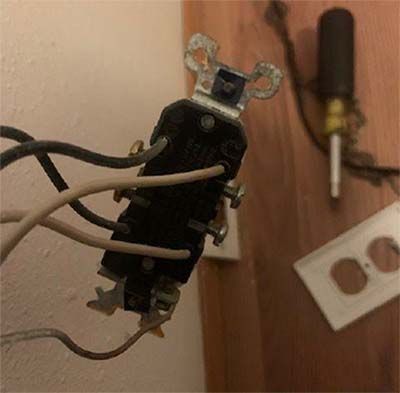 A wall electrical outlet hanging from wires, with a screwdriver and cover plate nearby on a wooden surface.