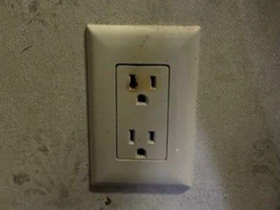 A slightly discolored, two-slot wall electrical outlet set against a plain, light-colored wall.