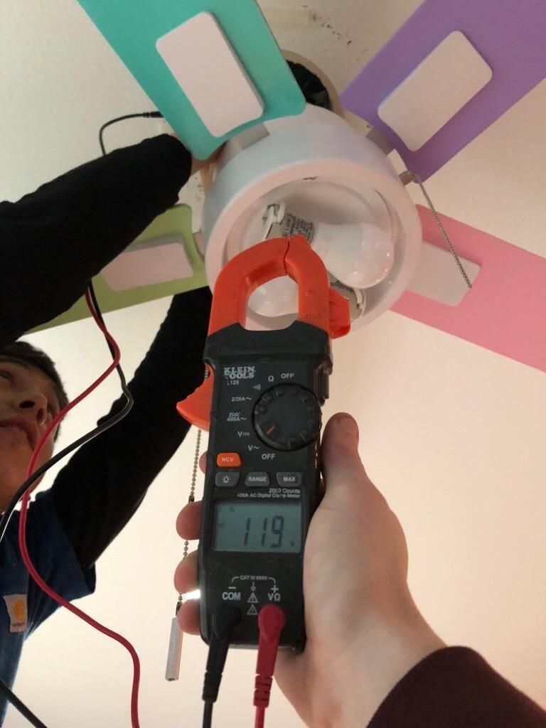 Person using a multimeter to test a ceiling light with colored fan blades on a pink ceiling.