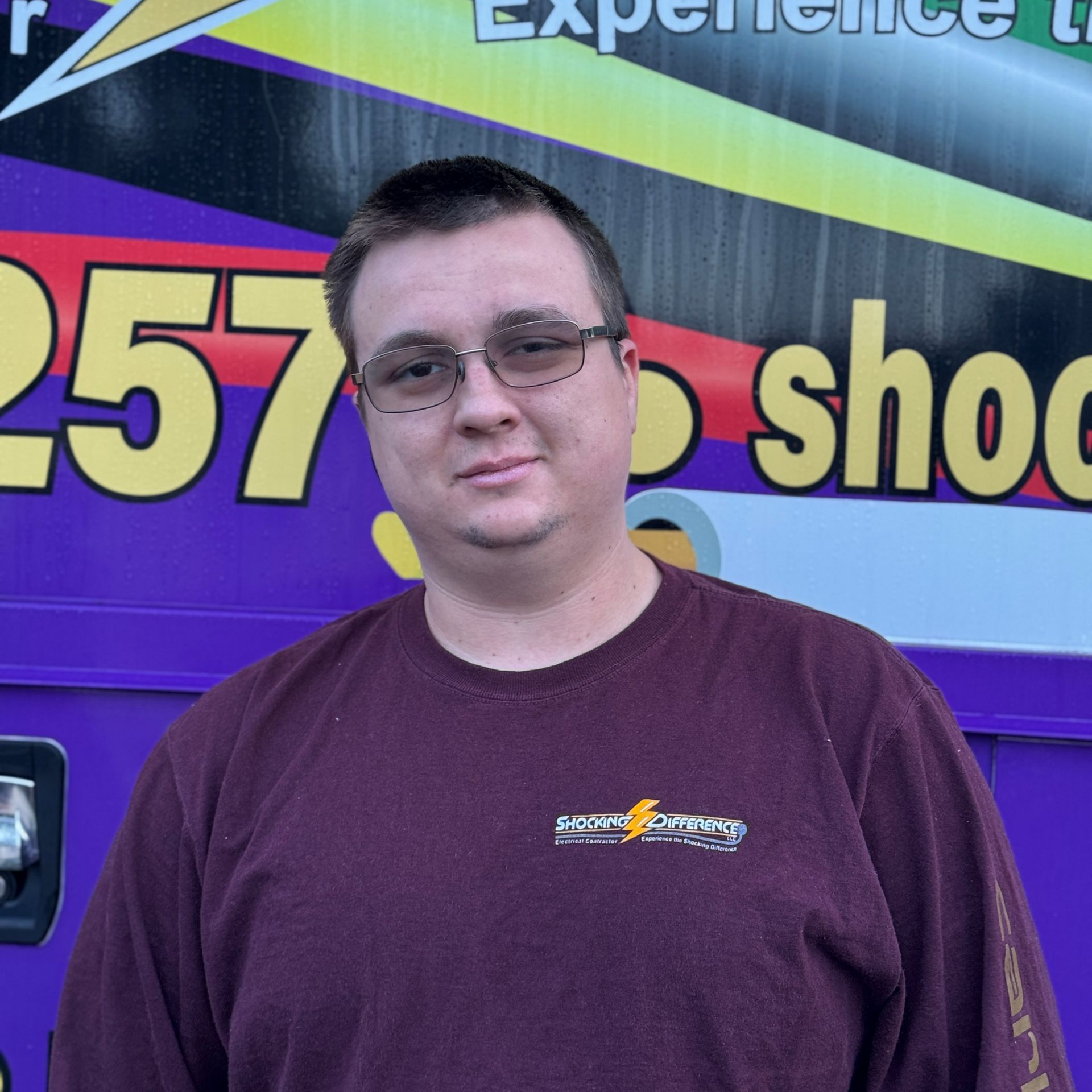 Man standing in front of a purple and yellow event banner, wearing glasses and a maroon shirt.