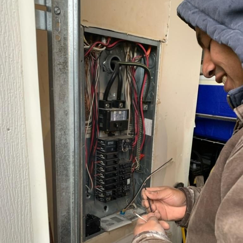 A person wearing a hooded jacket works on the exposed wiring inside an open electrical panel.