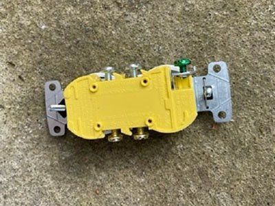 A yellow electrical wall outlet receptacle with silver metal mounting brackets and screws, set against a concrete surface.