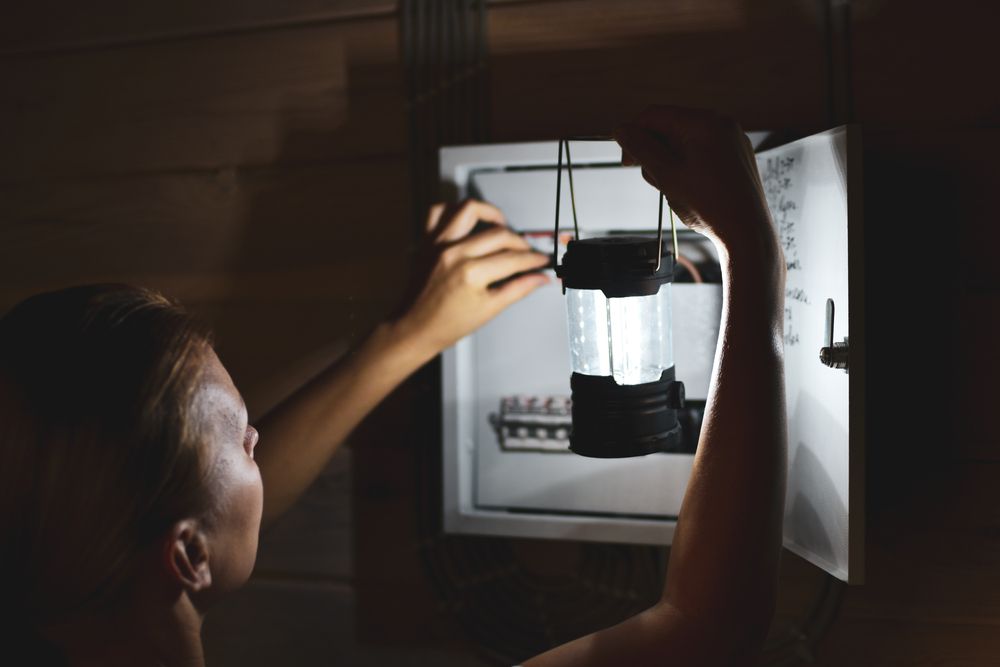 A person in a dim room illuminates an open electrical breaker box with a portable lantern.