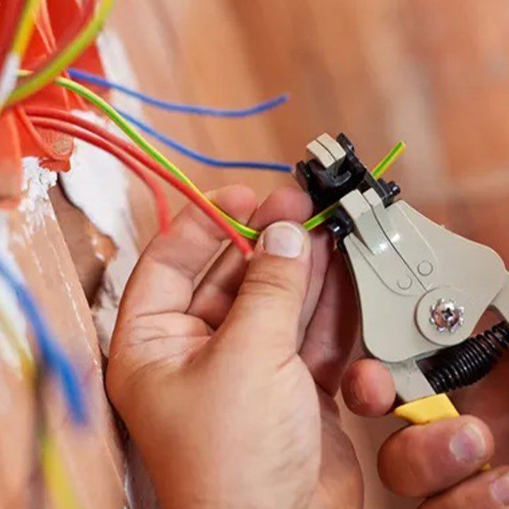 Hands using automatic wire strippers on a green and yellow electrical wire near a wall-mounted electrical junction box.
