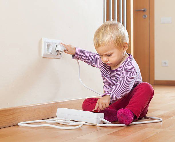 A toddler in purple stripes touches a power strip while plugging a cord into a wall outlet, creating an electrical hazard.