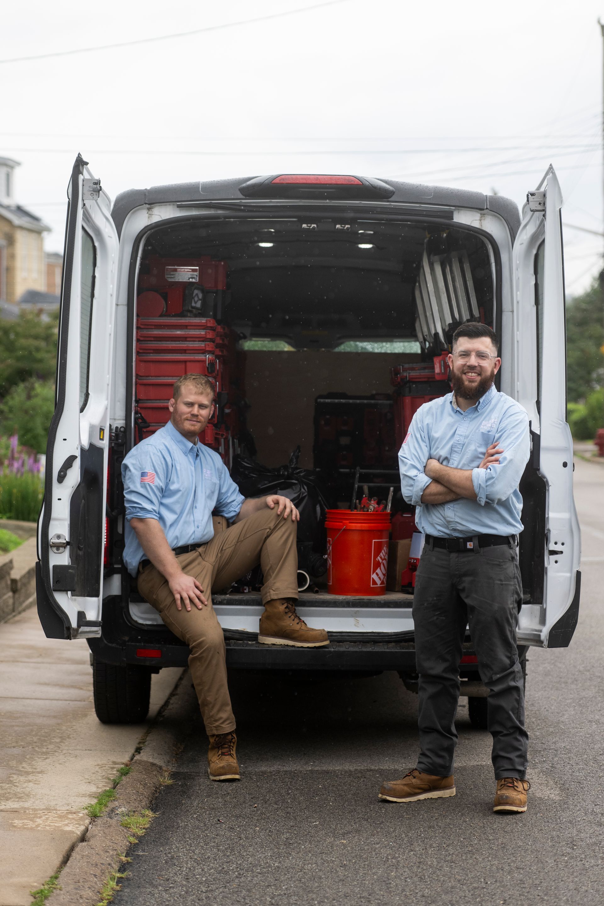 Two men stand by a white van with open doors, filled with tools. One man sits, other has arms crossed.