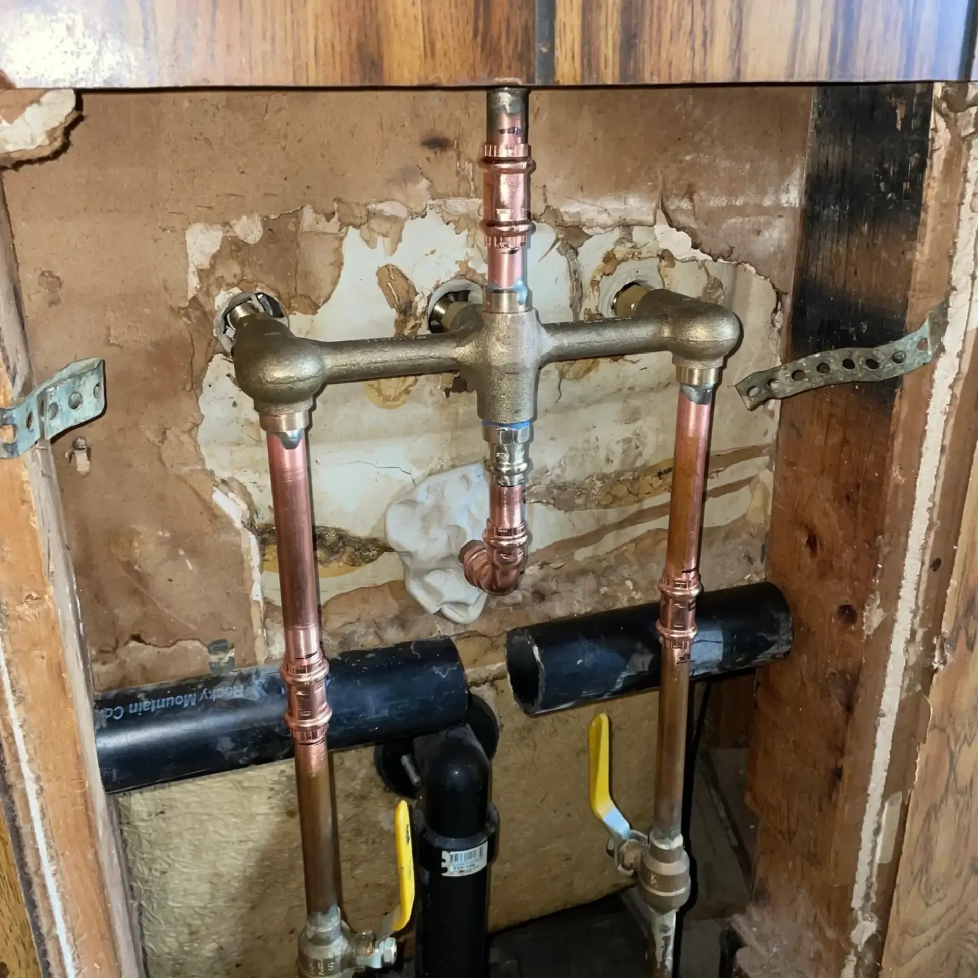 Copper pipes and brass fixtures with shutoff valves. Mounted on a damaged wall, with black pipe visible.