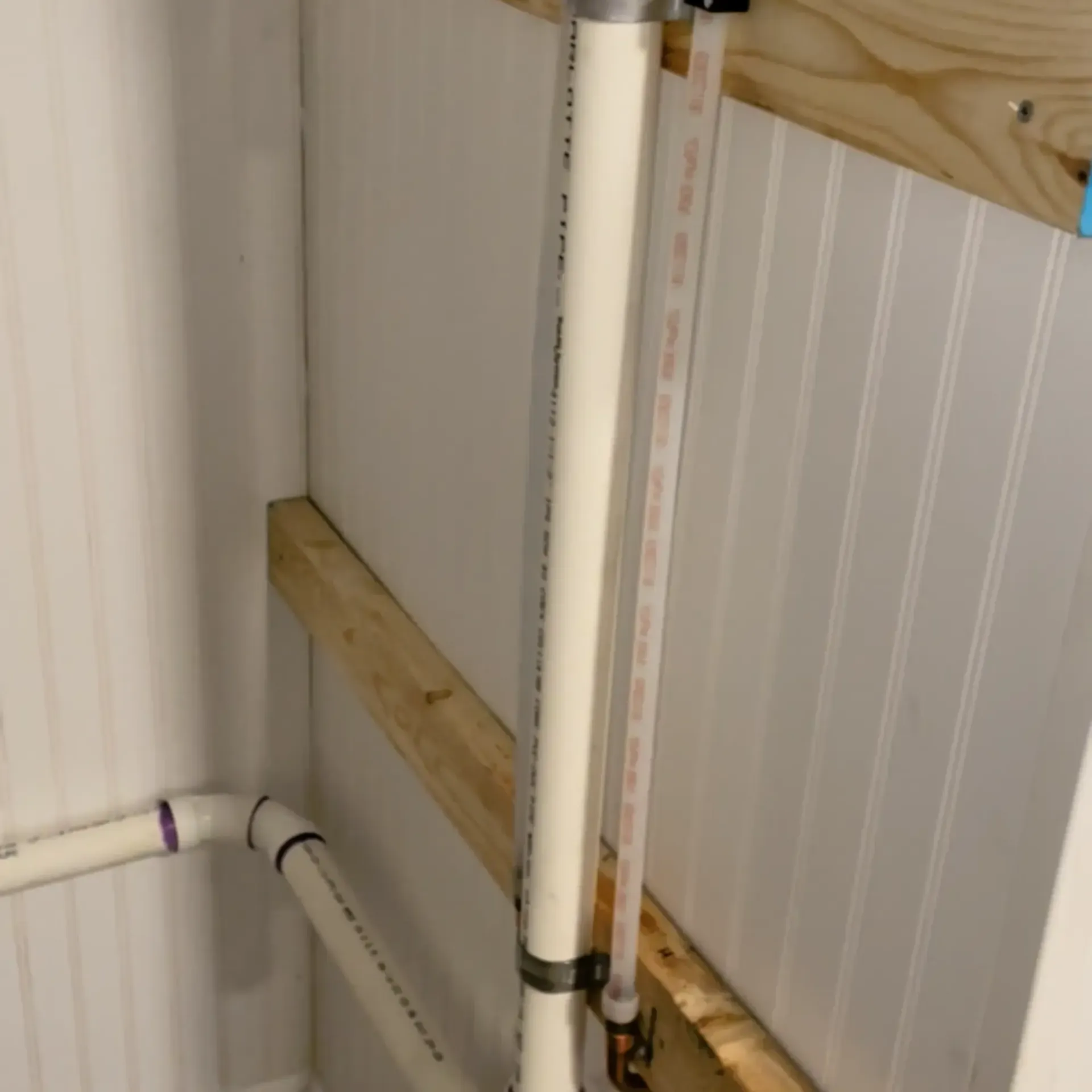 White PVC plumbing pipes against a white paneled wall with wooden support beams.