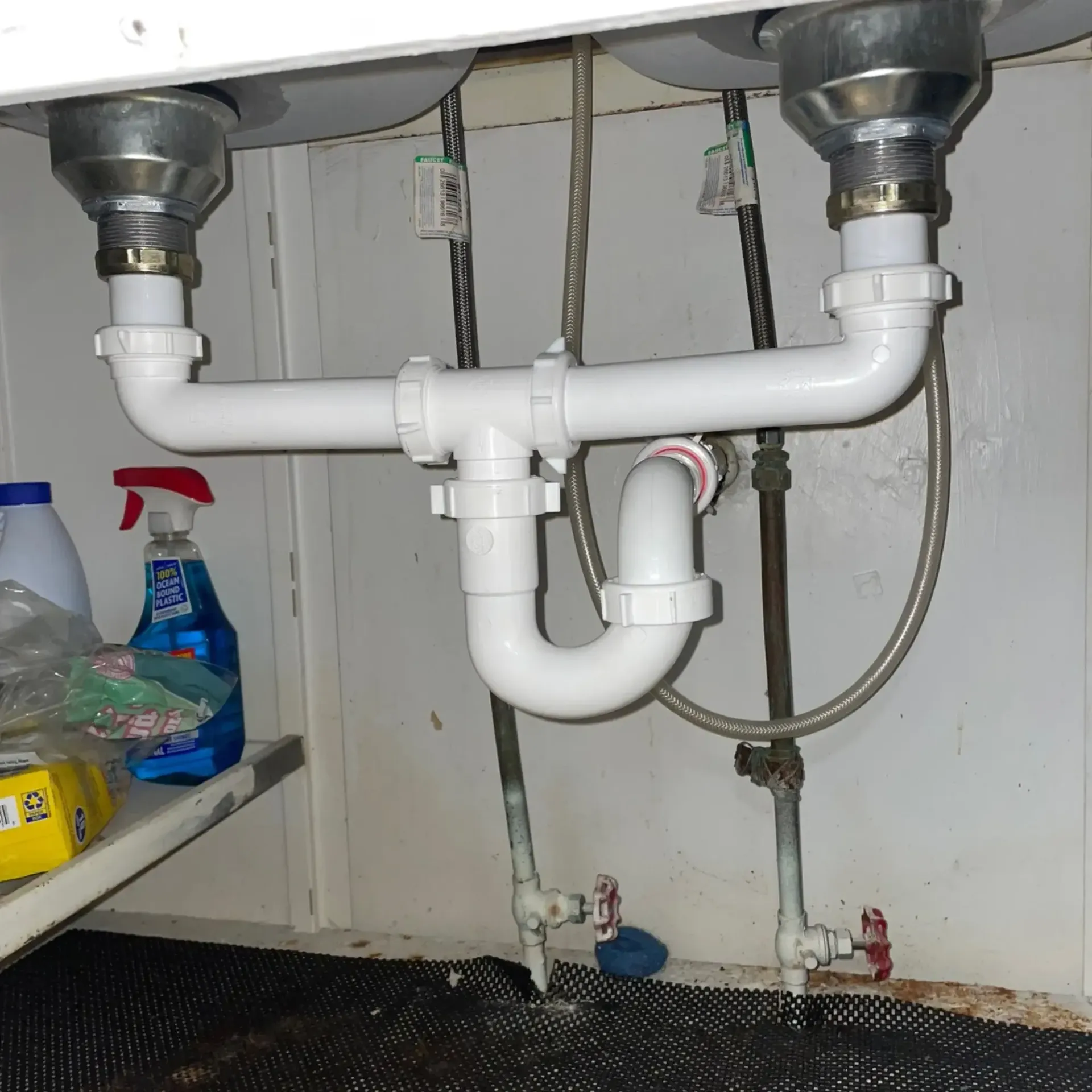 White plastic plumbing under a double sink in a white cabinet. Water valves are visible.