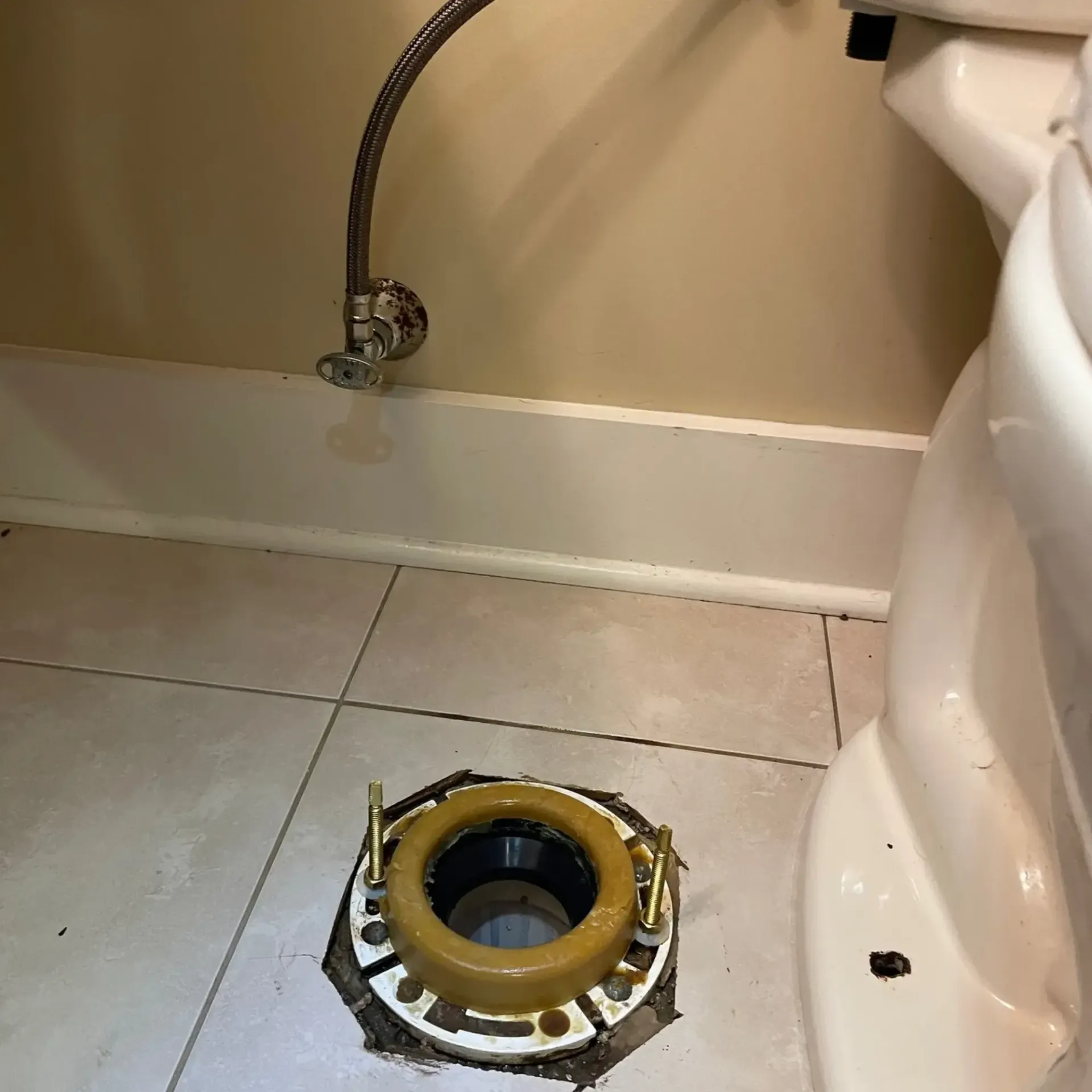 Toilet base with bolts, wax ring, and water supply line. White toilet bowl in a tiled bathroom.