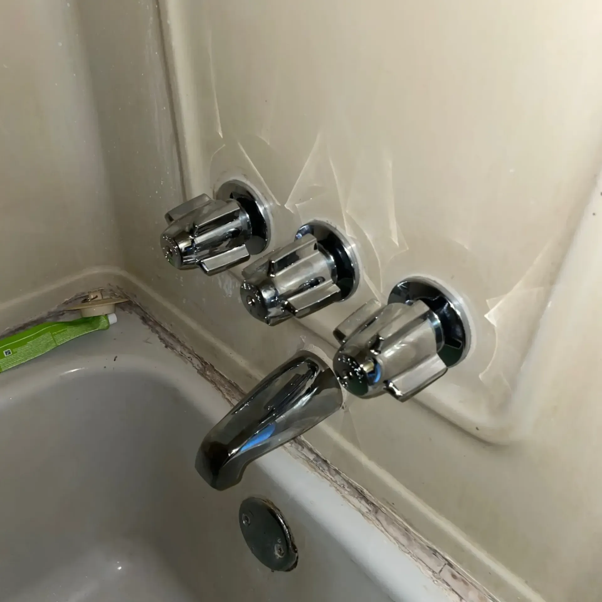 Bathtub faucet with three separate handles and a spout, mounted on a cream-colored wall.