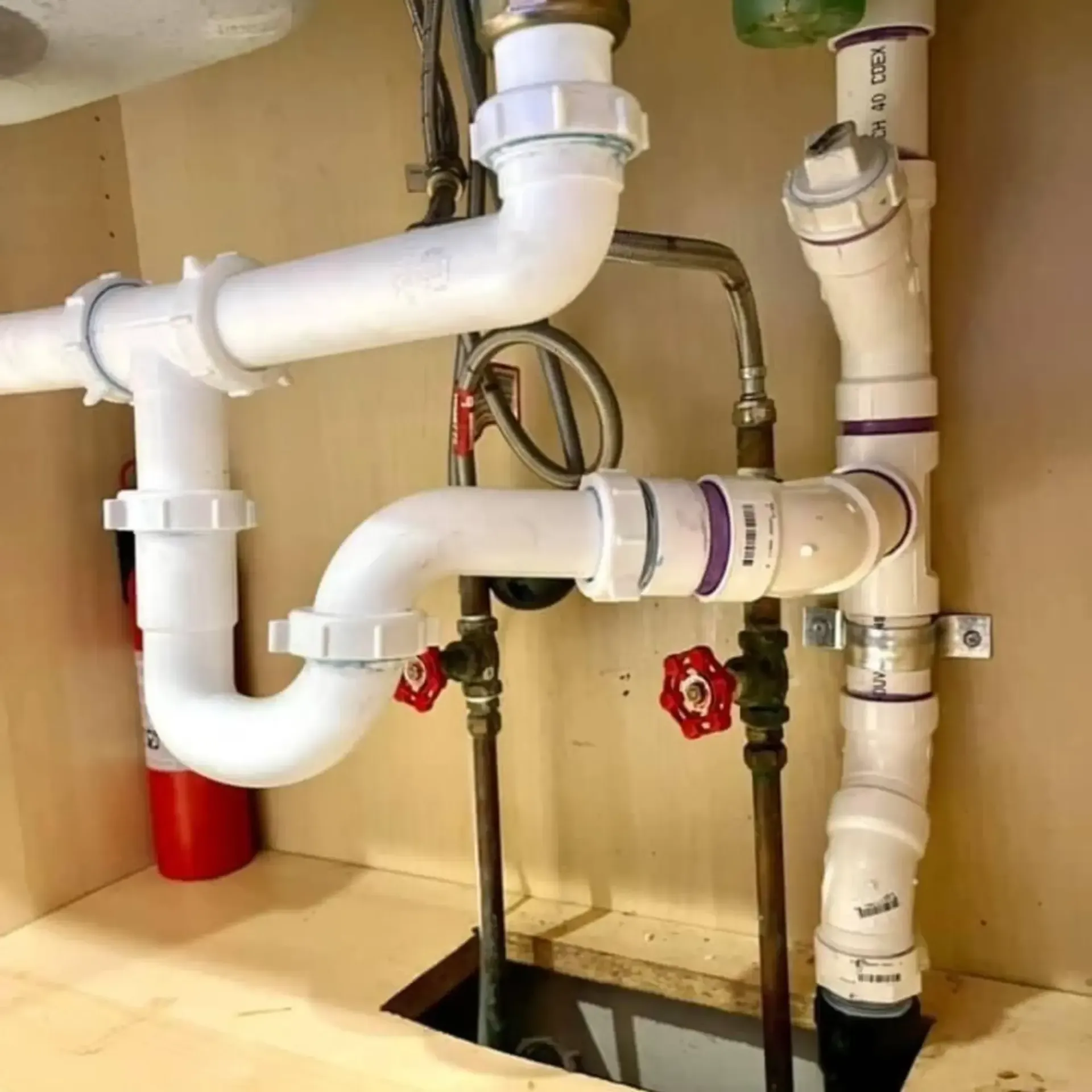 White PVC pipes connected under a sink. Plumbing includes multiple bends, joints, and drain connections with two red handled shut off valves.