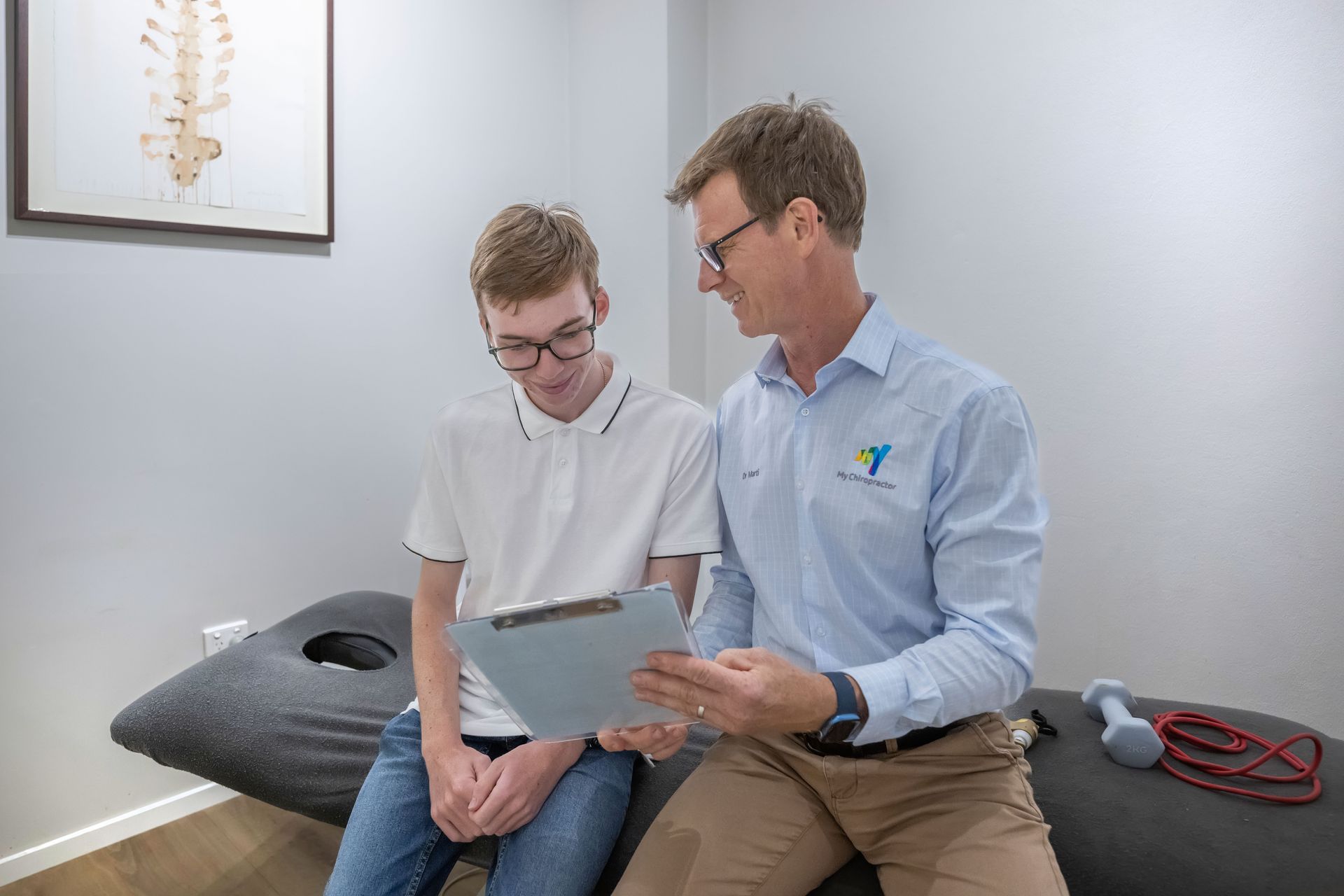 Two Men Sitting on An Exam Table, Looking at A Chart in A Clinical Office — My Chiropractor Mackay City in Mackay, QLD