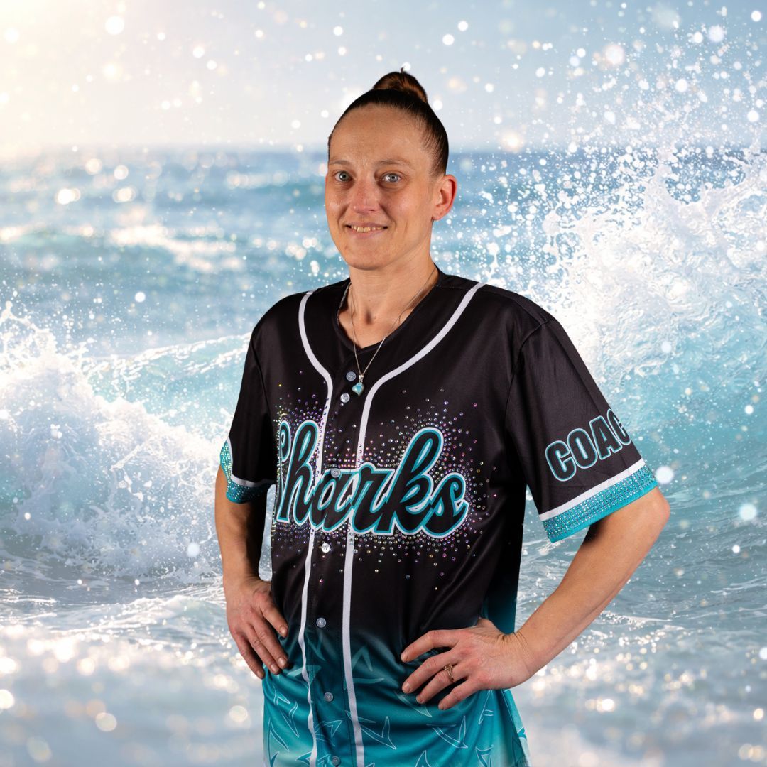 Woman in a Sharks baseball jersey smiles, hands on hips, against a background of ocean waves.