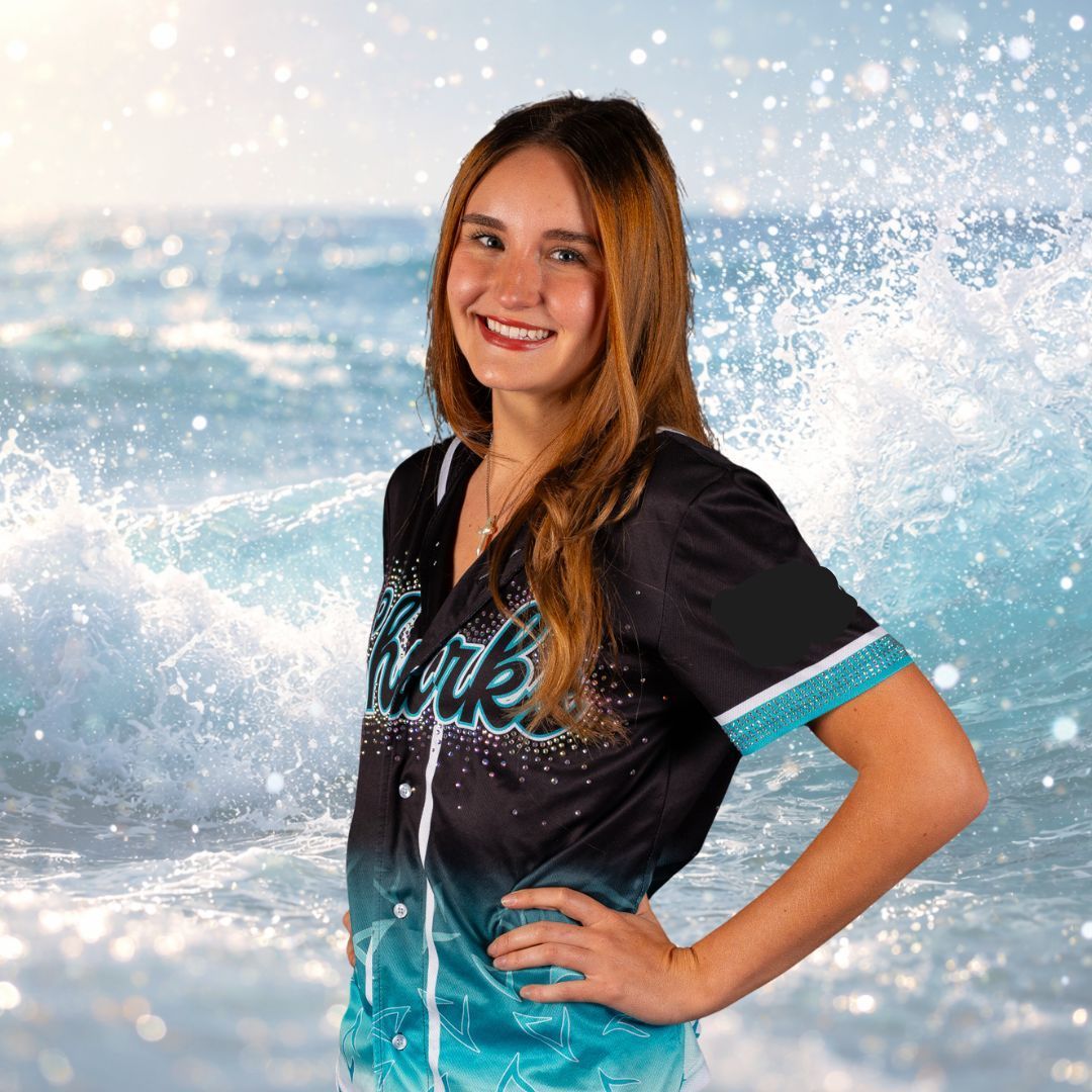 Woman in a baseball jersey smiles, hand on hip, against a backdrop of ocean waves.
