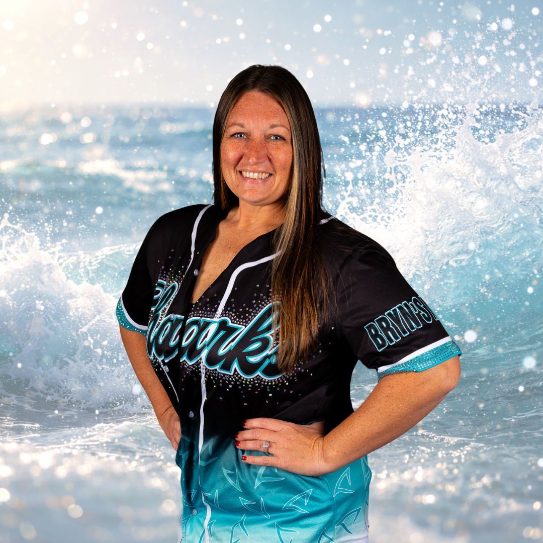 Woman in baseball jersey stands with hands on hips in front of ocean waves.