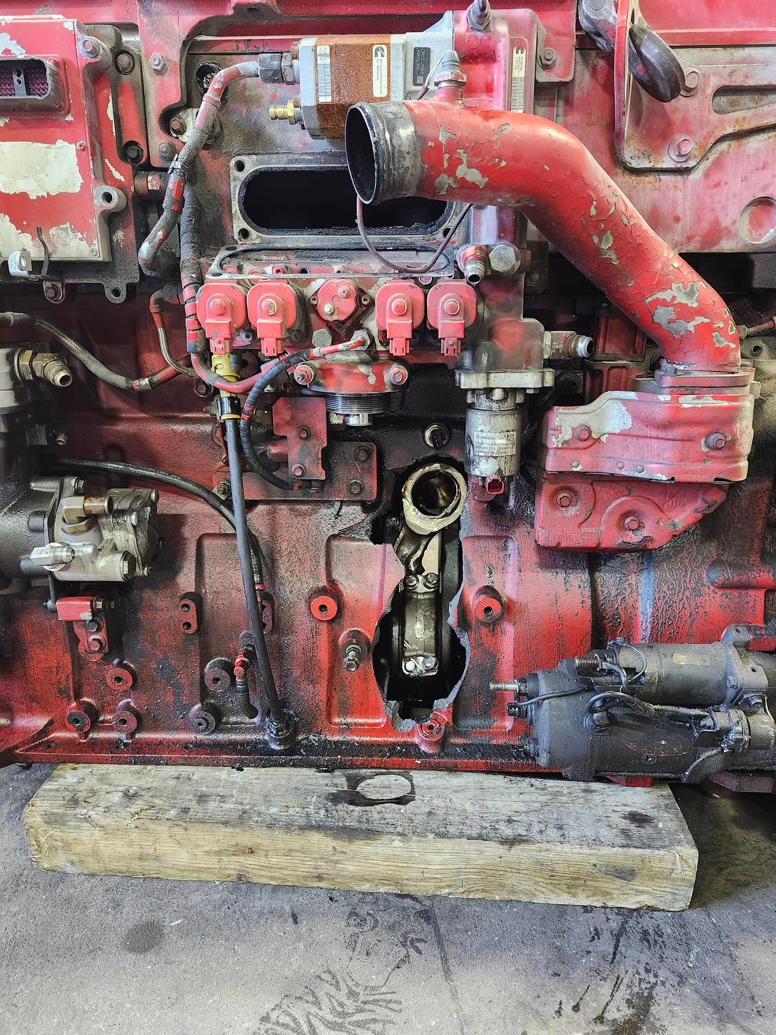 A damaged, red heavy-duty industrial engine with a large hole in the side of its engine block, resting on a wooden board.