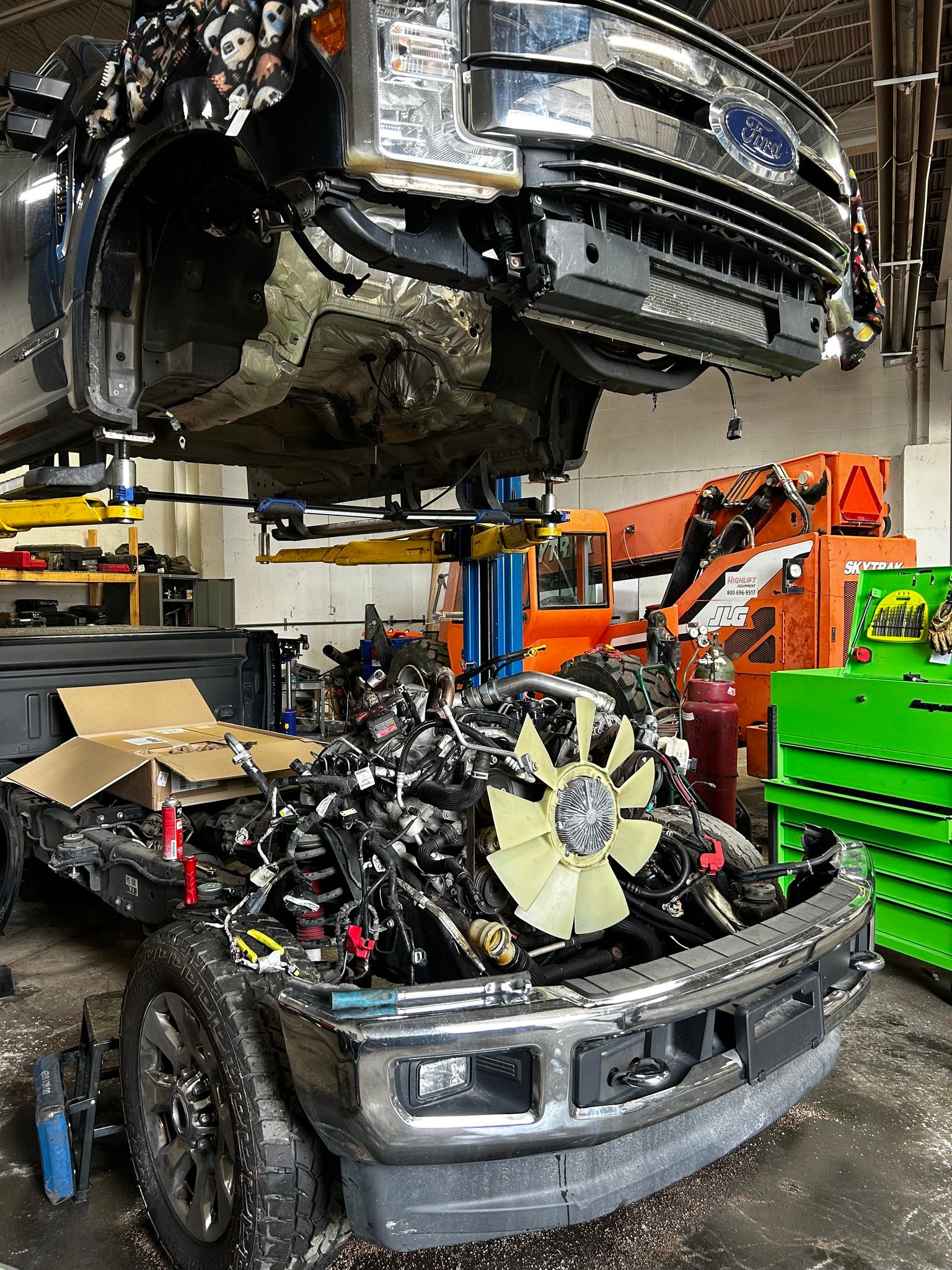 A truck cab is lifted high on a car lift in a repair shop, revealing the exposed engine and radiator frame below.