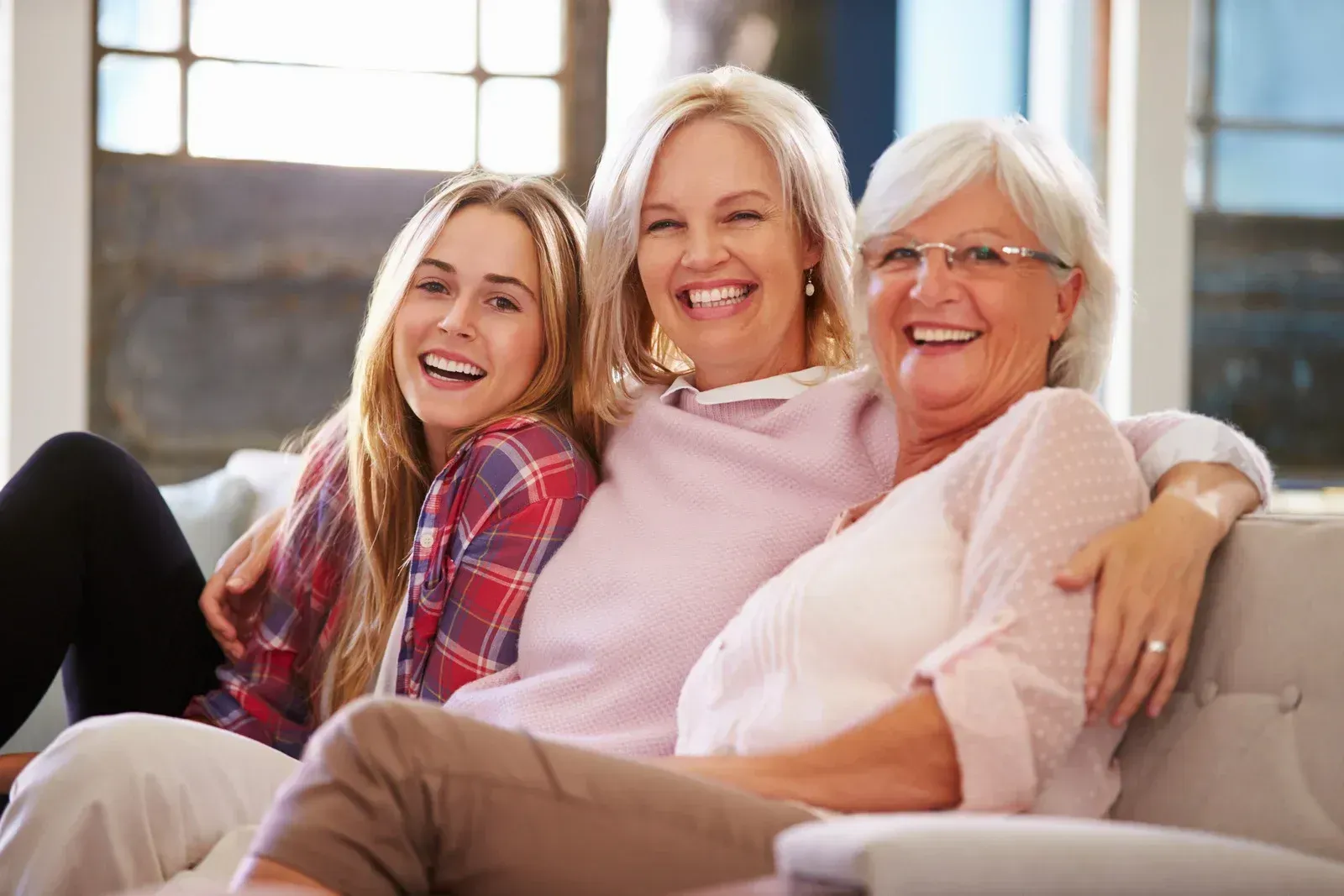Three smiling people sitting on a couch in a bright living room, with one in a plaid shirt and one in pink.