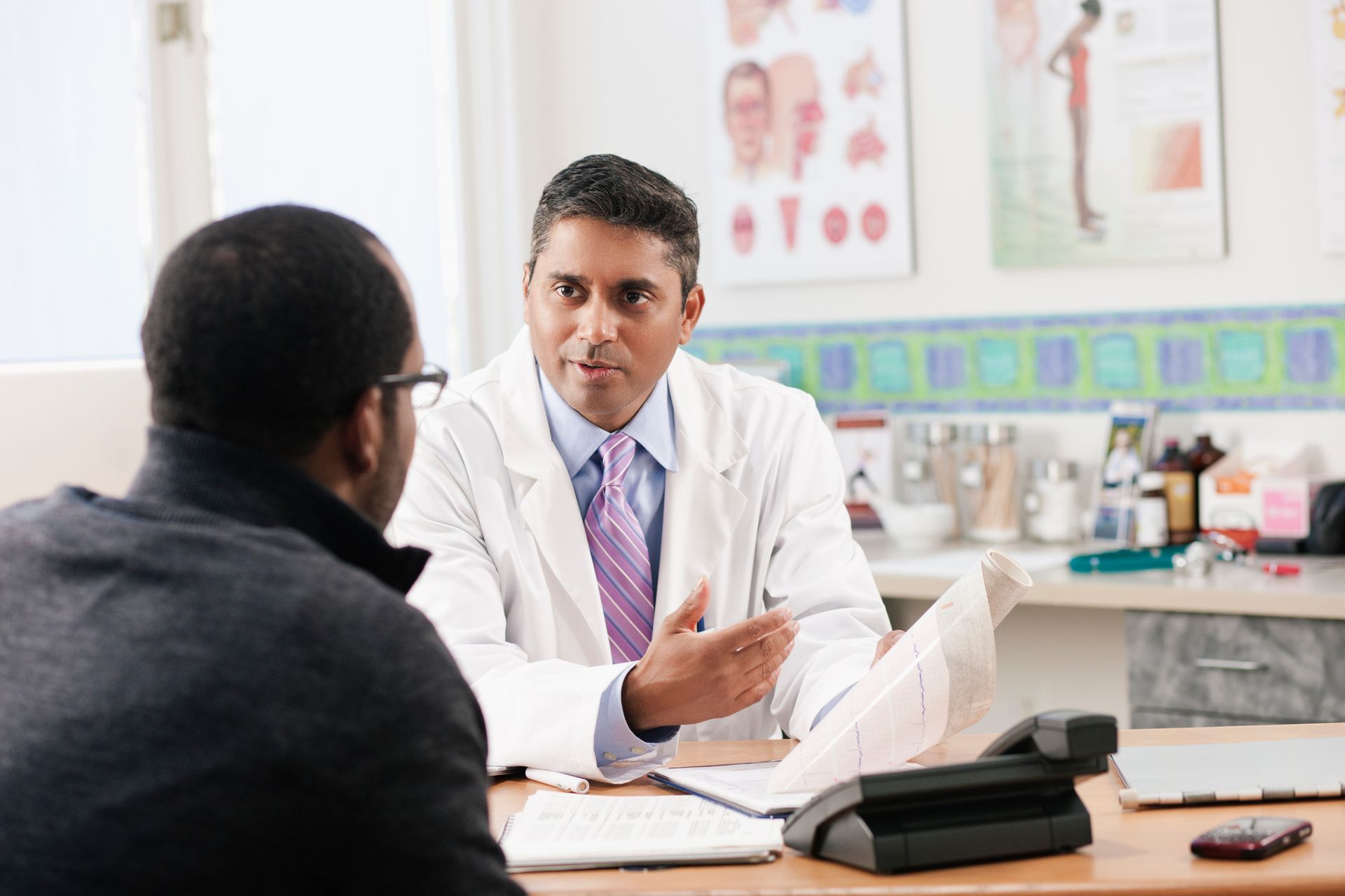 Doctor in a white coat speaking to a patient at a desk in a medical office.