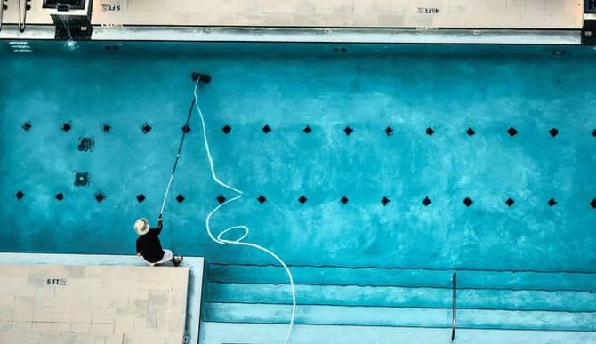 Person climbing a turquoise climbing wall with ropes and black holds beside a pool