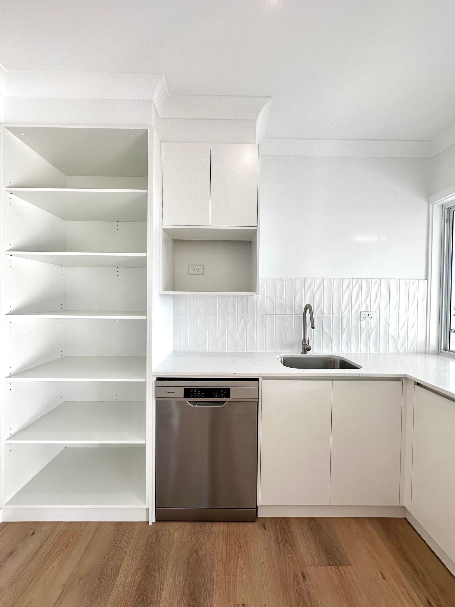 A kitchen with white cabinets , a stainless steel dishwasher , a sink , and shelves.