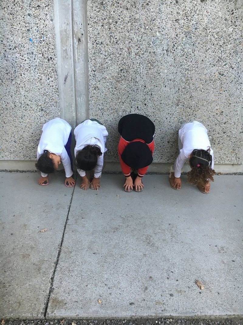 Three children are bending over on the sidewalk in front of a wall.