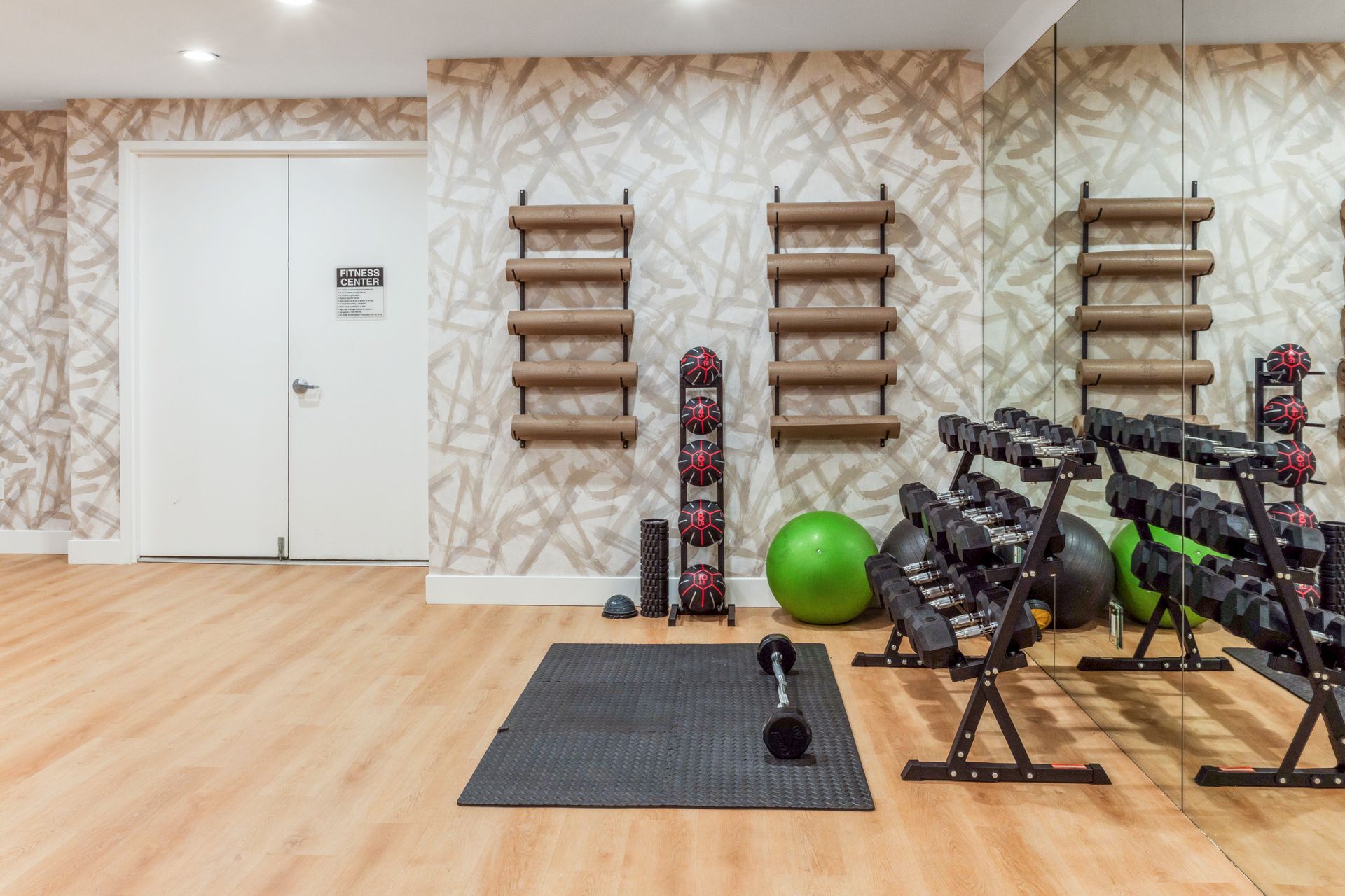 Bright gym studio with exercise mats, dumbbells, stability ball, and wall-mounted bars on a wood floor.
