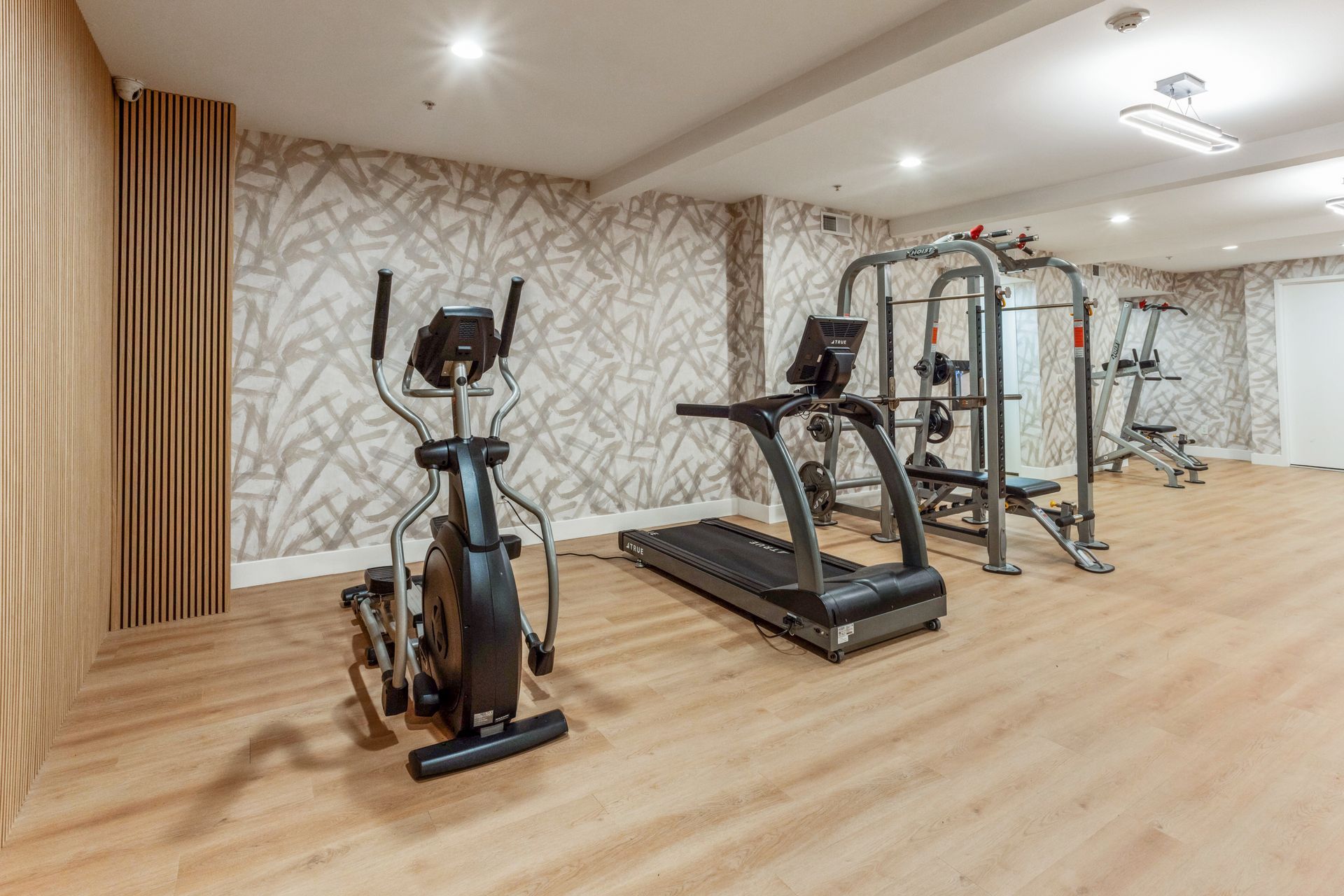 Empty home gym with two exercise machines, a treadmill, and mirrored wall on wood flooring.
