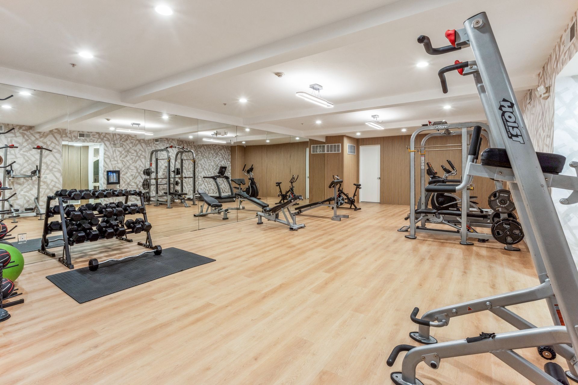 Spacious gym with weight machines, dumbbells, benches, and light wood flooring under bright ceiling lights