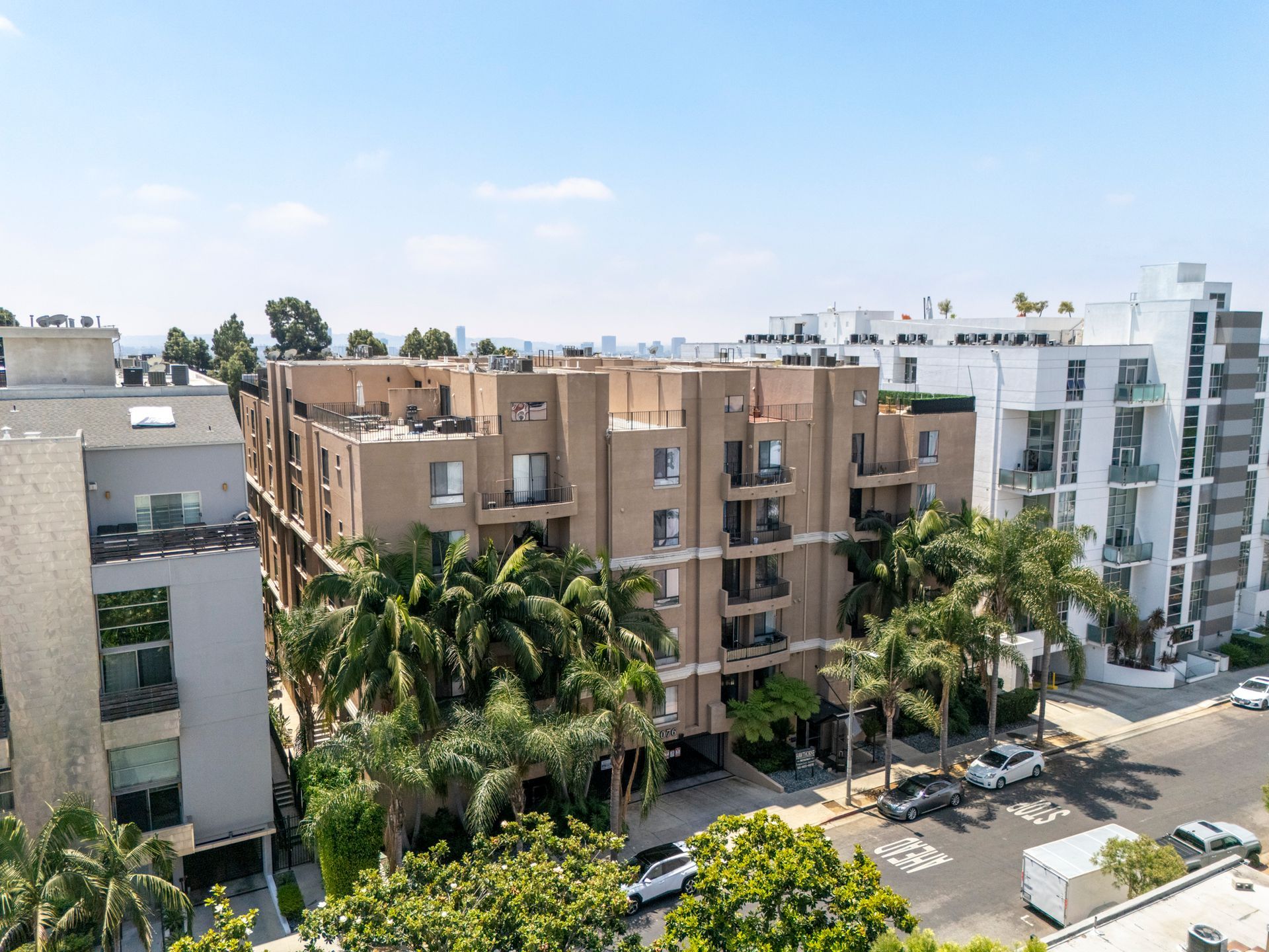 Urban street with tan apartment buildings, palm trees, and parked cars under a bright blue sky